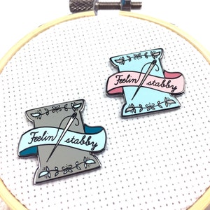 May include: Two enamel pins shaped like spools of thread with a needle and the text "Feelin' stabby" on a banner. The spool on the left is grey with a teal banner. The spool on the right is light blue with a pink banner.