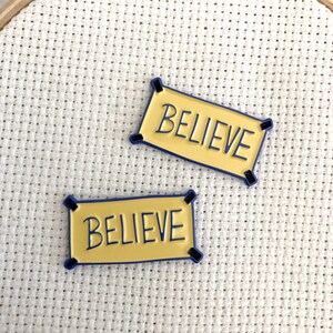K&ouml;nnte beinhalten: Zwei gelbe Emaille-Pins mit blauem Rand. Jeder Pin zeigt ein Banner mit dem Wort "BELIEVE" in Schwarz.