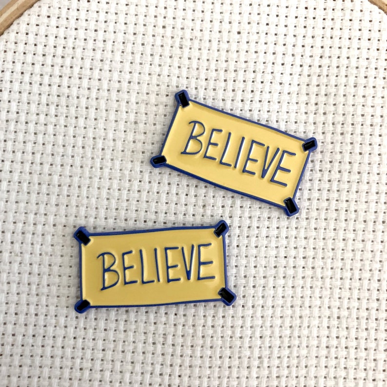 K&ouml;nnte beinhalten: Zwei gelbe Emaille-Pins mit blauem Rand. Jeder Pin zeigt ein Banner mit dem Wort "BELIEVE" in Schwarz.