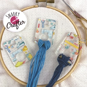 May include: Three clear plastic needle threaders with colorful designs and blue embroidery floss. The threaders are on a white fabric background.