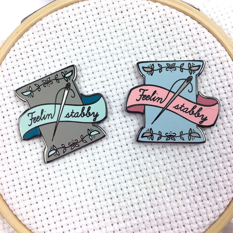 May include: Two enamel pins shaped like spools of thread with a needle and thread going through them. The spools are in different colours, one is grey and the other is blue. Both spools have a banner that says "Feelin' stabby".