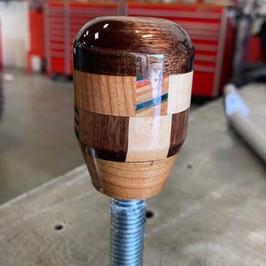 Könnte beinhalten: Handgefertigter Schaltknauf aus Holz mit glänzendem Finish. Der Knauf hat ein Mosaikdesign mit abwechselnd hellen und dunklen Holzsegmenten und ist auf einer silbernen Schraube montiert. Die Oberseite ist dunkleres, poliertes Holz.