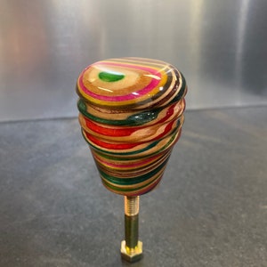 Könnte beinhalten: Ein handgefertigter, mehrfarbiger Holzknauf mit einer Messingschraube. Der Knauf besteht aus Schichten von Holz in den Farben Rot, Grün, Pink und Braun. Die Oberseite ist abgerundet und poliert, wodurch die Holzmaserung sichtbar wird. Die Schraube ist ca. 5 cm lang.