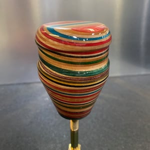 May include: A handcrafted wooden top with a colorful, layered design. The top features a rounded head and a tapered body, with a brass-colored bolt extending from the bottom. The wood layers display a variety of colors, including red, blue, green, and yellow.