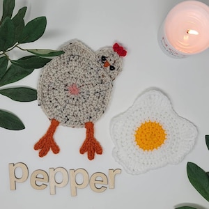 May include: Two crocheted pot holders, one shaped like a chicken and the other like a fried egg. The chicken pot holder is beige with brown legs and a red comb. The egg pot holder is white with a yellow yolk. The word "pepper" is written in wooden letters on a white surface.