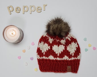 May include: Red knit hat with a white heart pattern and a brown faux fur pom pom. The hat has a small brown leather tag with the word "pepper" on it.