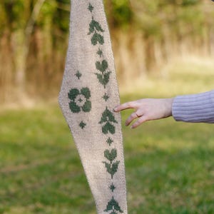 May include: A beige knitted scarf with a pointed end, held up against a blurred green background. The scarf features a repeating pattern of green floral and leaf motifs. The scarf is a long, narrow shape.