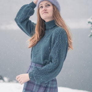 Peut inclure: Une femme portant un pull bleu en tricot avec un motif en tricot torsadé et une jupe en plaid violet. Elle porte un bonnet violet clair et se tient dans la neige.