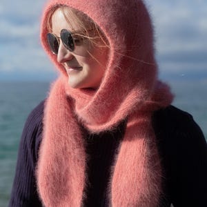 May include: A soft, fluffy, light pink scarf and hood set. The hood frames the face and neck, and the scarf drapes down the front. The set is made of a fuzzy material, creating a warm and cozy look. The person is wearing a dark blue sweater and sunglasses.