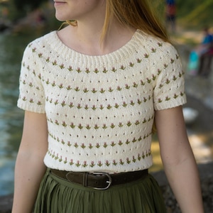 May include: Cream-coloured, short-sleeved sweater with a round neckline. The sweater features a repeating pattern of small pink and green floral motifs. A dark brown belt cinches the waist, paired with a long, olive green skirt.