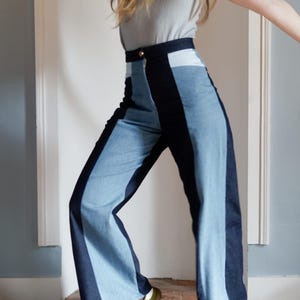 May include: High-waisted, wide-leg denim pants with a color-block design. The pants feature a light blue denim panel down the center of each leg, flanked by darker blue denim. A gold button secures the waistband. The model is wearing a gray t-shirt and green sneakers.