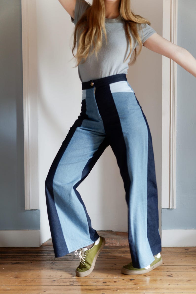 May include: High-waisted, wide-leg denim pants with a color-block design. The pants feature a light blue denim panel down the center of each leg, flanked by darker blue denim. A gold button secures the waistband. The model is wearing a gray t-shirt and green sneakers.