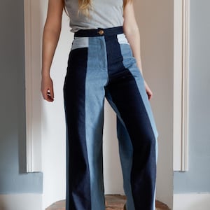 May include: High-waisted, wide-leg denim pants with a color-block design. The pants feature panels of light blue, medium blue, and dark blue denim. A gold button secures the waistband. The model is wearing a gray t-shirt and green sneakers.