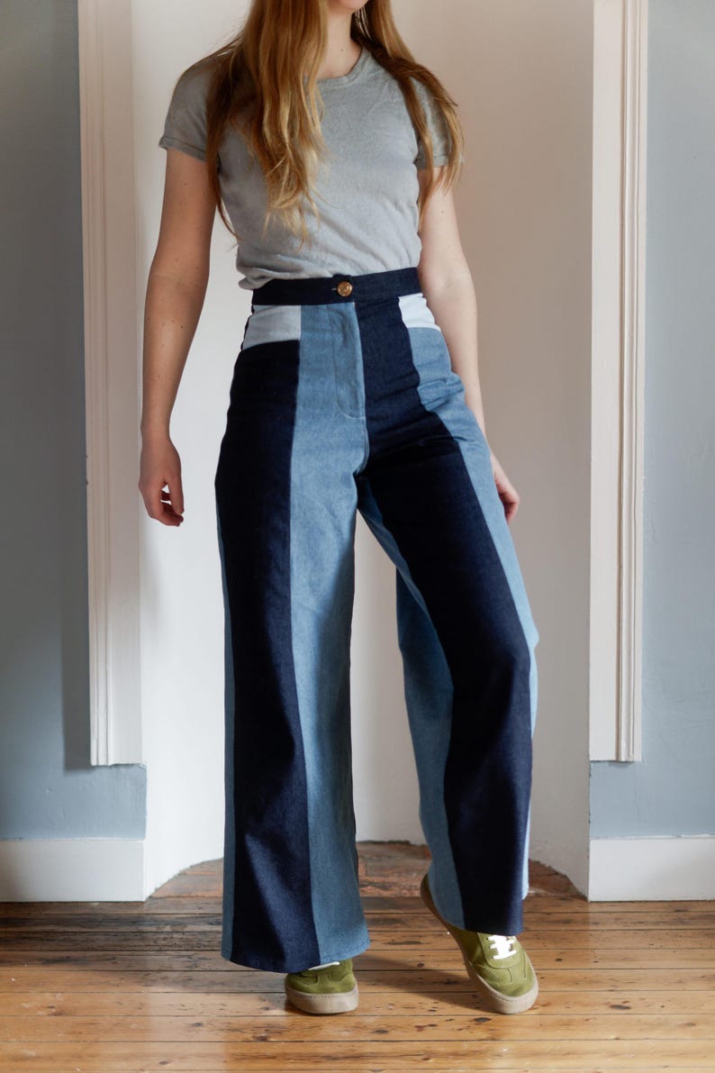 May include: High-waisted, wide-leg denim pants with a color-block design. The pants feature panels of light blue, medium blue, and dark blue denim. A gold button secures the waistband. The model is wearing a gray t-shirt and green sneakers.