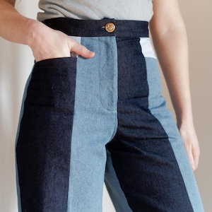 May include: Two-tone denim pants with a high waist and a button closure. The pants feature a dark blue and light blue color block design. The model is wearing a gray t-shirt. The pants have pockets.