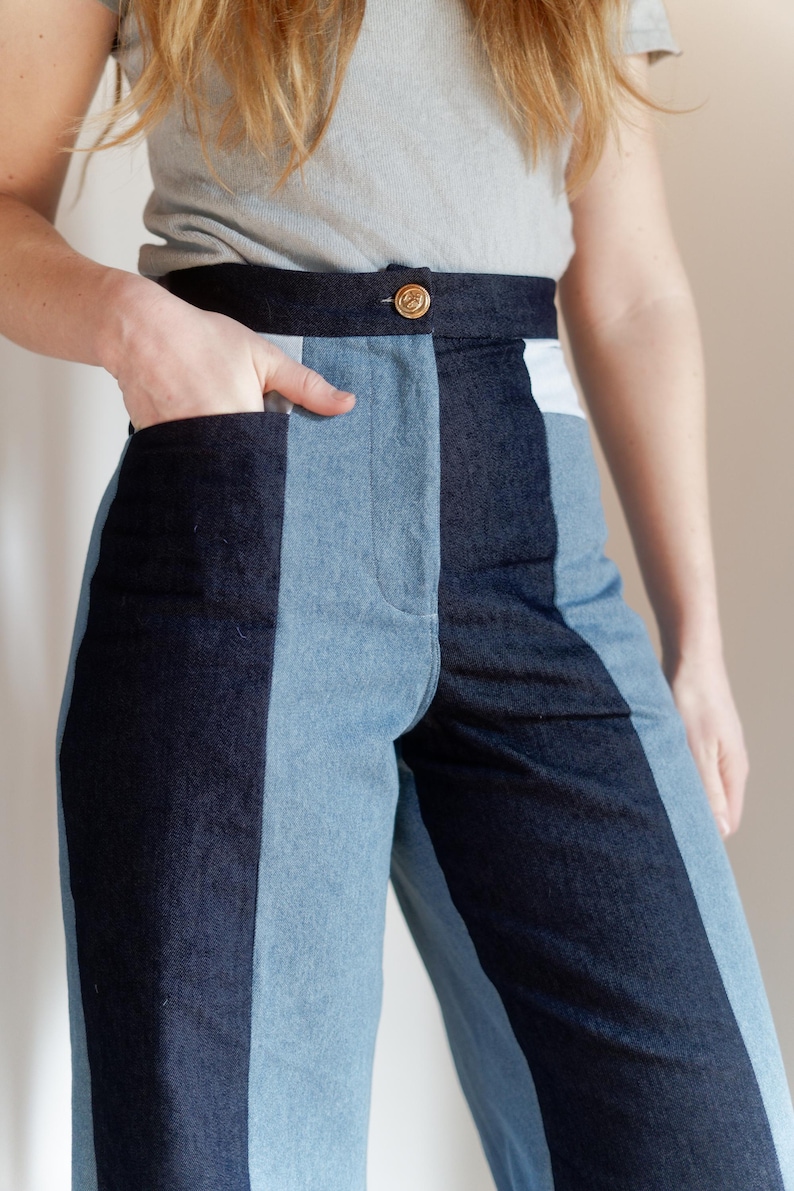 May include: Two-tone denim pants with a high waist and a button closure. The pants feature a dark blue and light blue color block design. The model is wearing a gray t-shirt. The pants have pockets.
