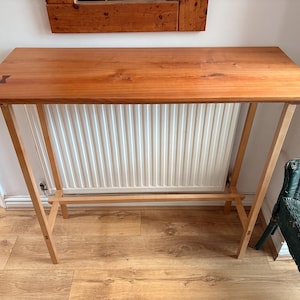 Puede incluir: Una mesa consola de madera con una tapa rectangular y patas en ángulo. La tapa es de color marrón cálido y las patas son de un tono de madera más claro. La mesa mide aproximadamente 122 cm de largo y 41 cm de ancho.