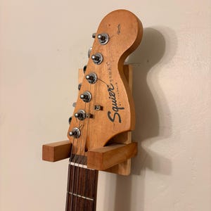 Puede incluir: Un soporte de pared de madera para guitarra que sostiene un clavijero de guitarra Squier Strat. El clavijero es de color marrón claro con texto negro y clavijas plateadas. El diapasón es marrón oscuro.