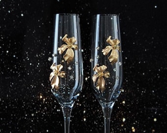 May include: Two clear glass champagne flutes with gold floral embellishments. The flutes are on a black background.
