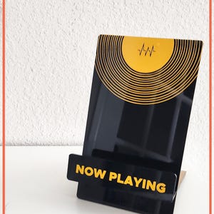 May include: Black acrylic record display stand with a yellow and black vinyl record design. The stand features the words "NOW PLAYING" in yellow. The stand is designed to hold a vinyl record.