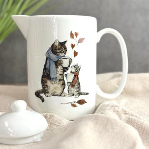 May include: White ceramic pitcher with a cat illustration. The design features a mother cat and kitten wearing scarves and holding mugs, with brown heart-shaped leaves. A white lid sits in front of the pitcher.