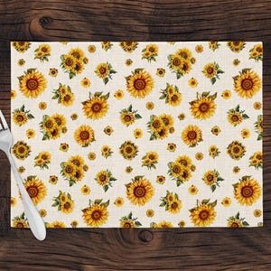 May include: A rectangular placemat featuring a repeating pattern of yellow sunflowers with brown centers and green leaves on a light beige background, accompanied by a single silver-toned metal fork positioned on the left side of the mat.