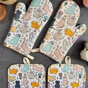 May include: Set of two white oven mitts and two pot holders with a colorful cat pattern. The pattern includes cats of different colors, sizes, and poses, along with flowers, hearts, and the word "Meow".