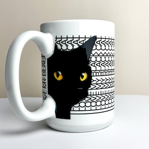 May include: White ceramic mug featuring a black cat design with yellow eyes. The mug has a large white handle and a decorative pattern. Text on the mug reads "WE MY BLACK CAT".