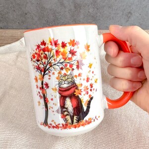 May include: White ceramic mug with an orange handle and rim. The mug features a cat wearing a red scarf, sitting under a tree with autumn leaves. The mug is held by a hand.