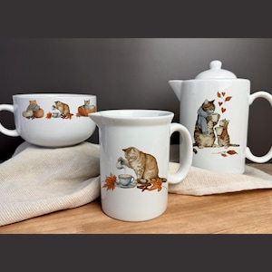 May include: A white ceramic tea set featuring cat illustrations. The set includes a large mug, a creamer, and a teapot with a lid. The cat designs are in shades of brown and orange, with autumn leaves. The set is on a wooden surface.