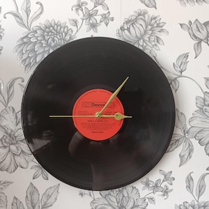 May include: A circular wall clock crafted from a black vinyl record featuring a central red label with the text Chevron and various printed details. The clock has three thin, gold-coloured metal hands positioned at the centre of the record.