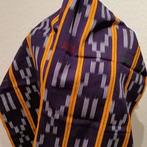K&ouml;nnte beinhalten: Ein violettes Textil mit wei&szlig;em und orangefarbenem geometrischem Muster. Das Textil ist &uuml;ber einer Holzform drapiert. Es hat einen Fransenrand. Das Wort "JUM" ist rot gestickt. Wahrscheinlich ein Schal oder Tuch.