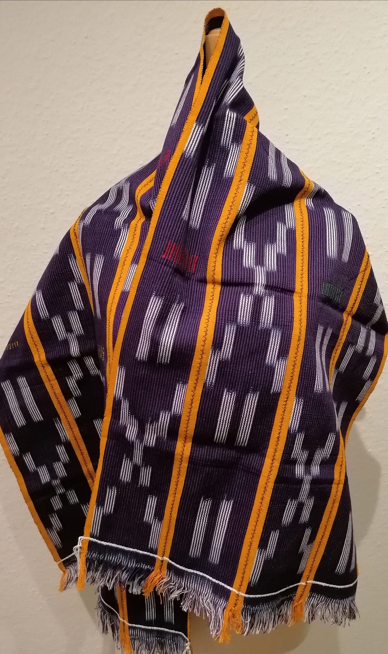 K&ouml;nnte beinhalten: Ein violettes Textil mit wei&szlig;em und orangefarbenem geometrischem Muster. Das Textil ist &uuml;ber einer Holzform drapiert. Es hat einen Fransenrand. Das Wort "JUM" ist rot gestickt. Wahrscheinlich ein Schal oder Tuch.