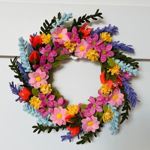 May include: A felt flower wreath with pink, yellow, orange, and purple flowers. The wreath is decorated with green leaves and blue sprigs.