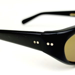 May include: Black plastic sunglasses with brown lenses and gold accents. The sunglasses have a rounded shape and a thin frame.