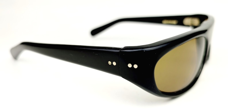May include: Black plastic sunglasses with brown lenses and gold accents. The sunglasses have a rounded shape and a thin frame.