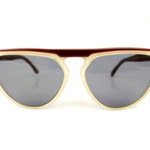 May include: Sunglasses with a cream-colored frame and dark gray lenses. The top bar is a deep burgundy color. The sunglasses have a retro, semi-rimless design.