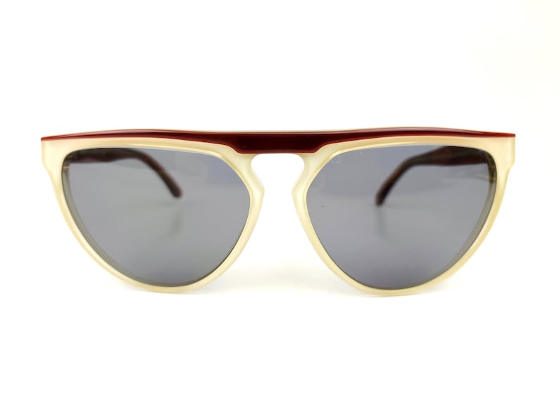 May include: Sunglasses with a cream-colored frame and dark gray lenses. The top bar is a deep burgundy color. The sunglasses have a retro, semi-rimless design.