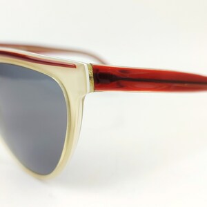 May include: Stylish sunglasses with a cream-colored frame and dark gray lenses. The temples are a rich red-brown color. The sunglasses have a retro, oversized design.