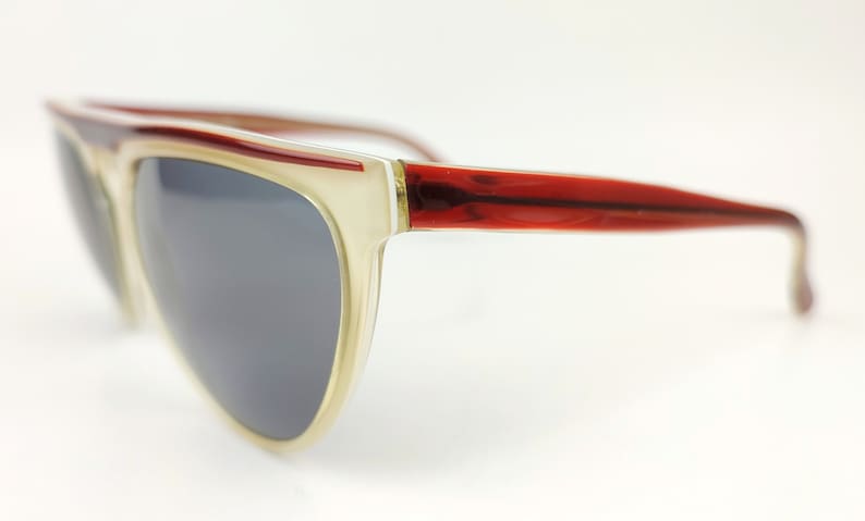 May include: Stylish sunglasses with a cream-colored frame and dark gray lenses. The temples are a rich red-brown color. The sunglasses have a retro, oversized design.