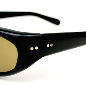 May include: Black plastic sunglasses with brown lenses. The sunglasses have a rounded shape and a thin frame.