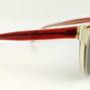 May include: Sunglasses with a rectangular frame. The frame is a light beige color with a red stripe along the top and red arms. The lenses are a dark gray color.