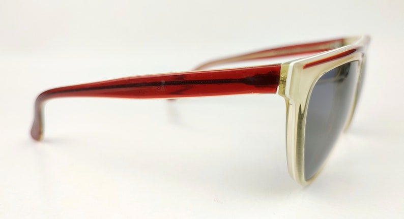 May include: Sunglasses with a rectangular frame. The frame is a light beige color with a red stripe along the top and red arms. The lenses are a dark gray color.