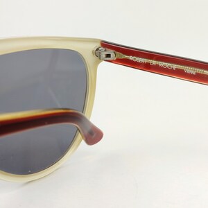 May include: Close-up of vintage sunglasses with a cream-colored frame and dark gray lenses. The temples are a gradient of red and brown, with the text "ROBERT LA ROCHE VIENNE" printed on the right temple. The sunglasses have a retro aesthetic.