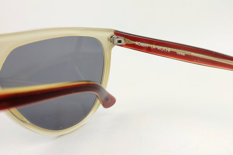 May include: Close-up of vintage sunglasses with a cream-colored frame and dark gray lenses. The temples are a gradient of red and brown, with the text "ROBERT LA ROCHE VIENNE" printed on the right temple. The sunglasses have a retro aesthetic.