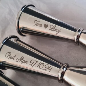 May include: Two stainless steel jiggers with engraved text, one reading "Tom ♥ Lucy", the other "Best Man 27.10.94". They are on a white surface. These are likely bar tools or gifts, with a polished, reflective finish.