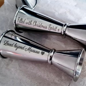 Op de afbeelding: Twee zilveren maatbekers met gegraveerde berichten. De ene beker heeft de tekst "Filled with Christmas Spirit x" en de andere "Loved beyond Measure".