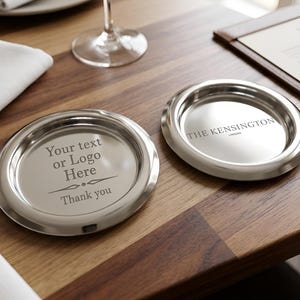 Könnte beinhalten: Zwei silberfarbene, runde Untersetzer mit erhöhtem Rand liegen auf einem Holztisch. Auf einem Untersetzer steht der Text "Your text or Logo Here" und "Thank you". Auf dem anderen steht "THE KENSINGTON LONDON".