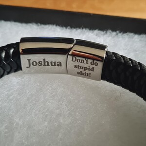 May include: Black braided leather bracelet with a silver clasp. The clasp is engraved with the name "Joshua" and the phrase "Don't do stupid shit!". The bracelet is presented in a black gift box with white padding.