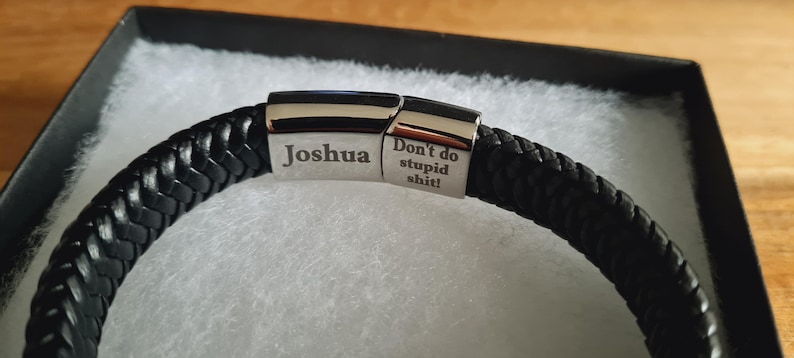 May include: Black braided leather bracelet with a silver clasp. The clasp is engraved with the name "Joshua" and the phrase "Don't do stupid shit!". The bracelet is presented in a black gift box with white padding.
